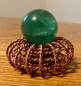 green crystal sphere on top of a braided copper base on a wooden surface.