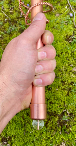 Hand holding a copper-colored tool with a clear crystal on a green moss background