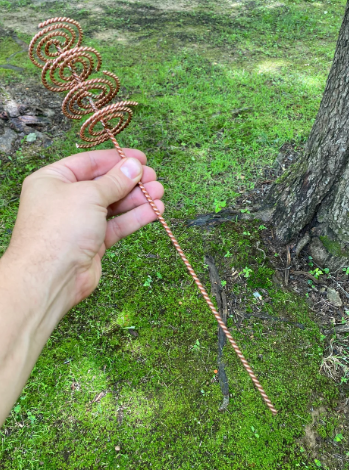 Hand holding a coiled copper tool against a natural background with grass and a tree.