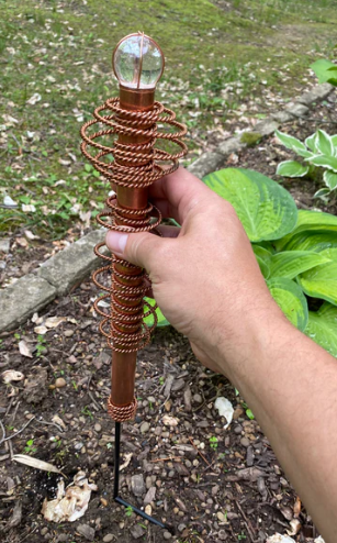 Hand holding a decorative metal spiral with a light bulb at the top, outdoors.