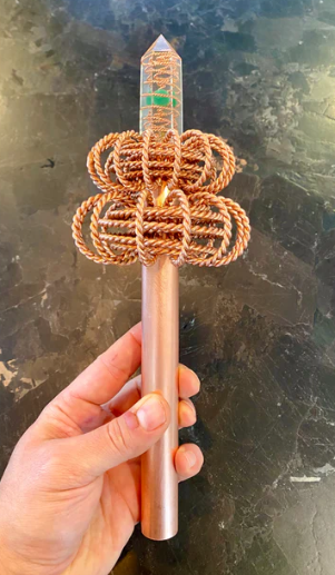 Hand holding a copper braided wand  with a decorative crystal and resin on a dark surface