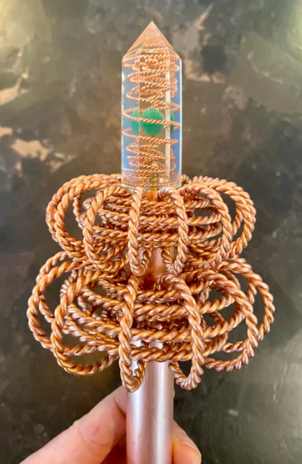 Crystal wand with braided copper design held by a hand on a dark surface