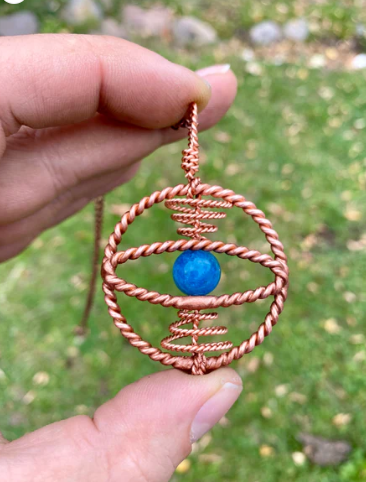 Hand holding a copper wire-wrapped pendant with a blue stone against a blurred natural background