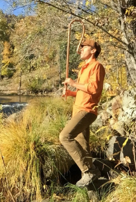 Person in an orange shirt and brown pants standing by a river with trees in the background
