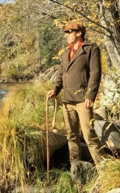 Man in outdoor setting with a cane, wearing a jacket and sunglasses, standing near a body of water.