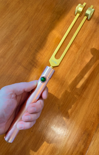 Hand holding a metallic tool with a green gemstone on a wooden surface