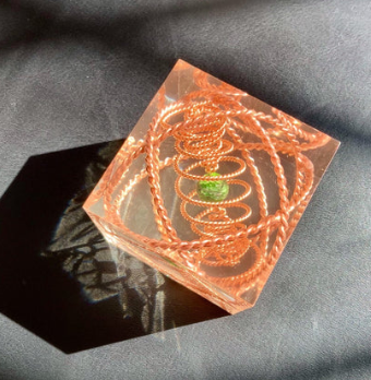 Decorative square object with copper spiral design and green gemstone on a dark fabric background